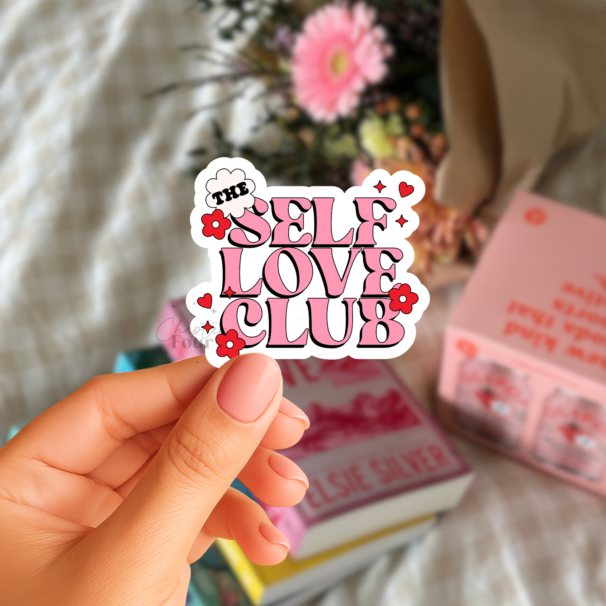 Hand holding a 'The Self Love Club' sticker with books and flowers in the background