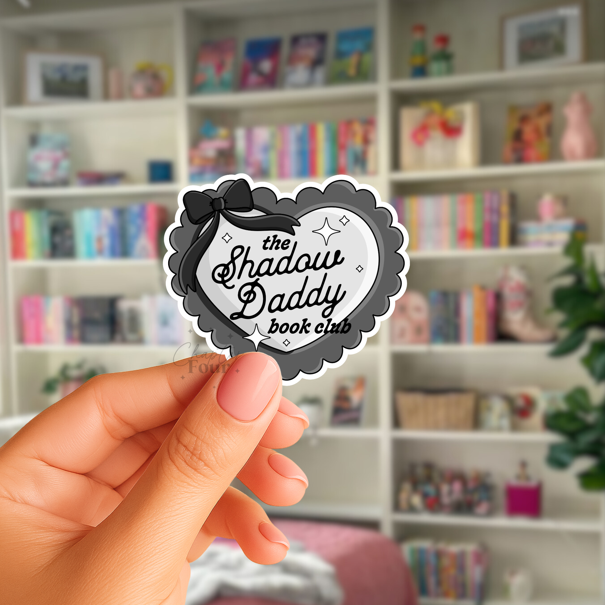 Hand holding a black and gray 'The Shadow Daddy Book Club' heart sticker in front of a bookshelf.