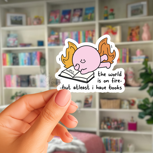 Hand holding a sticker with a cartoon character with 'The world is on fire but at least i have books' text in front of a bookshelf.