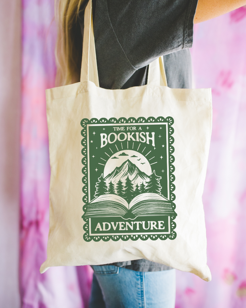 A person holding a beige tote bag with 'time for a bookish adventure' text printed on it, featuring a mountain and tree design.