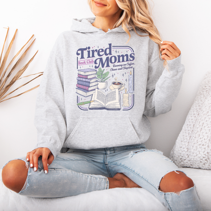 A woman wearing a light ash colored hoodie with a colorful graphic that reads 'Tired Moms Book Club' and showing ripped light-wash jeans, sitting on a white couch.