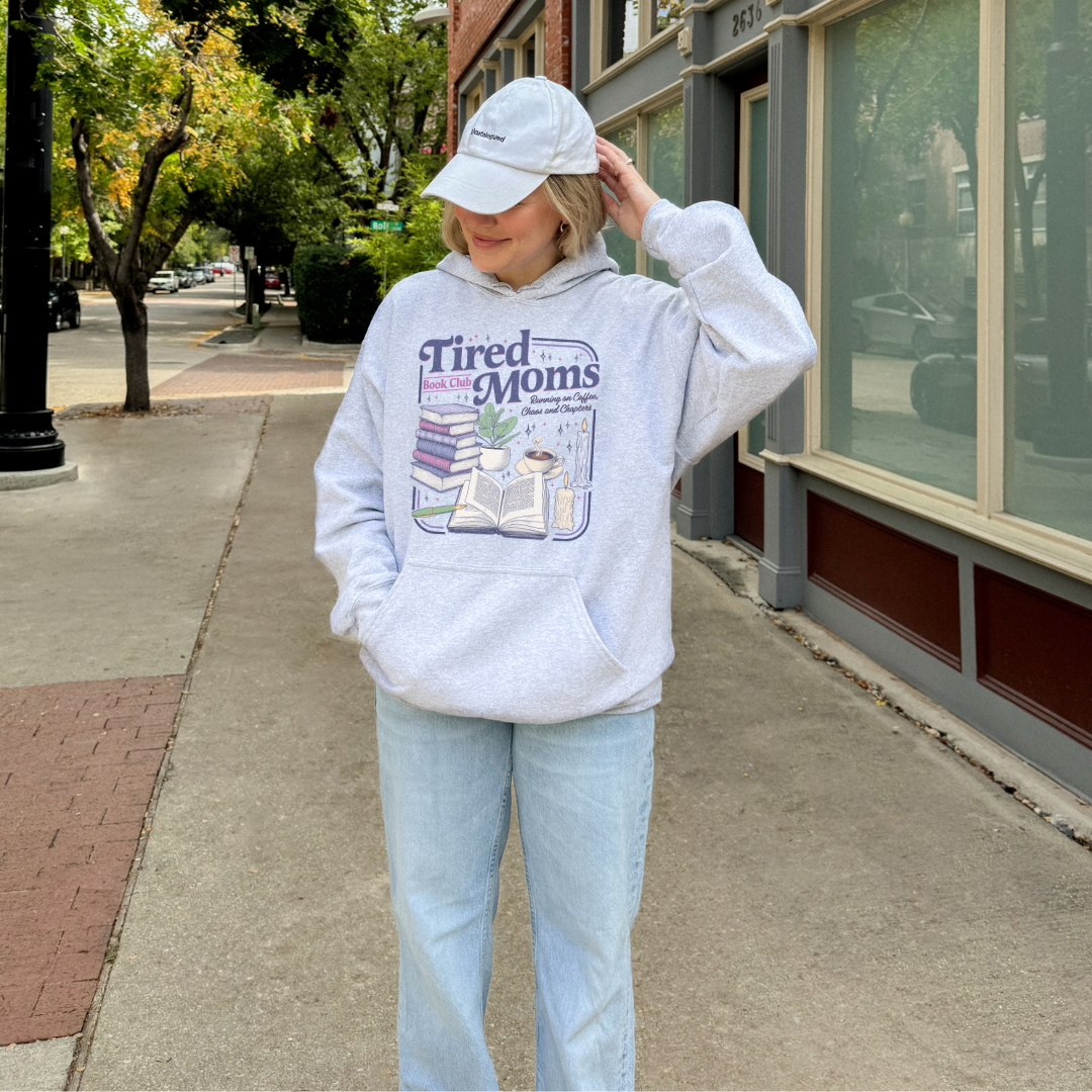 A woman stands on a city sidewalk wearing a ash colored oversized hoodie with a colorful graphic that reads 'Tired Moms Book Club' and a matching light gray baseball cap.