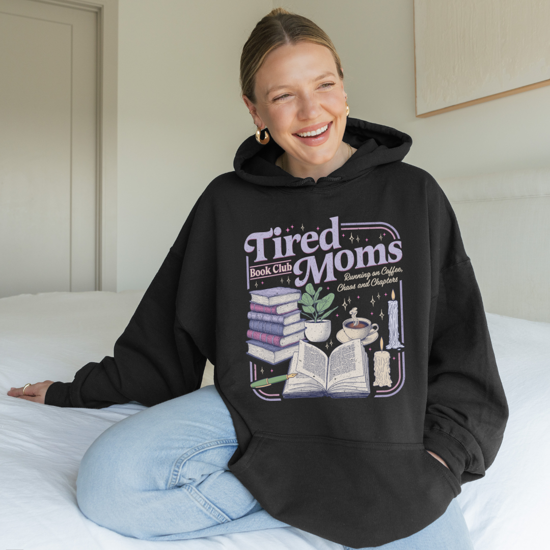 A woman sitting on a bed wearing a black oversized hoodie with a colorful graphic that reads 'Tired Moms Book Club' and showing light blue jeans in a bedroom setting.