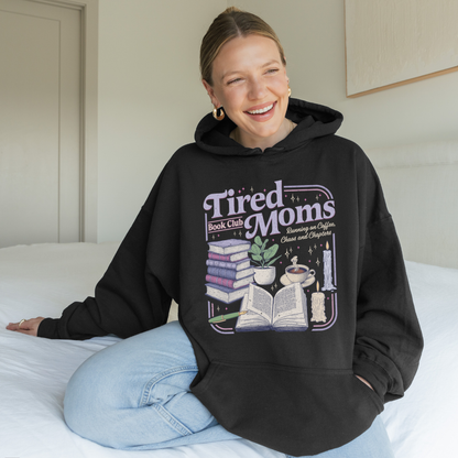 A woman sitting on a bed wearing a black oversized hoodie with a colorful graphic that reads 'Tired Moms Book Club' and showing light blue jeans in a bedroom setting.