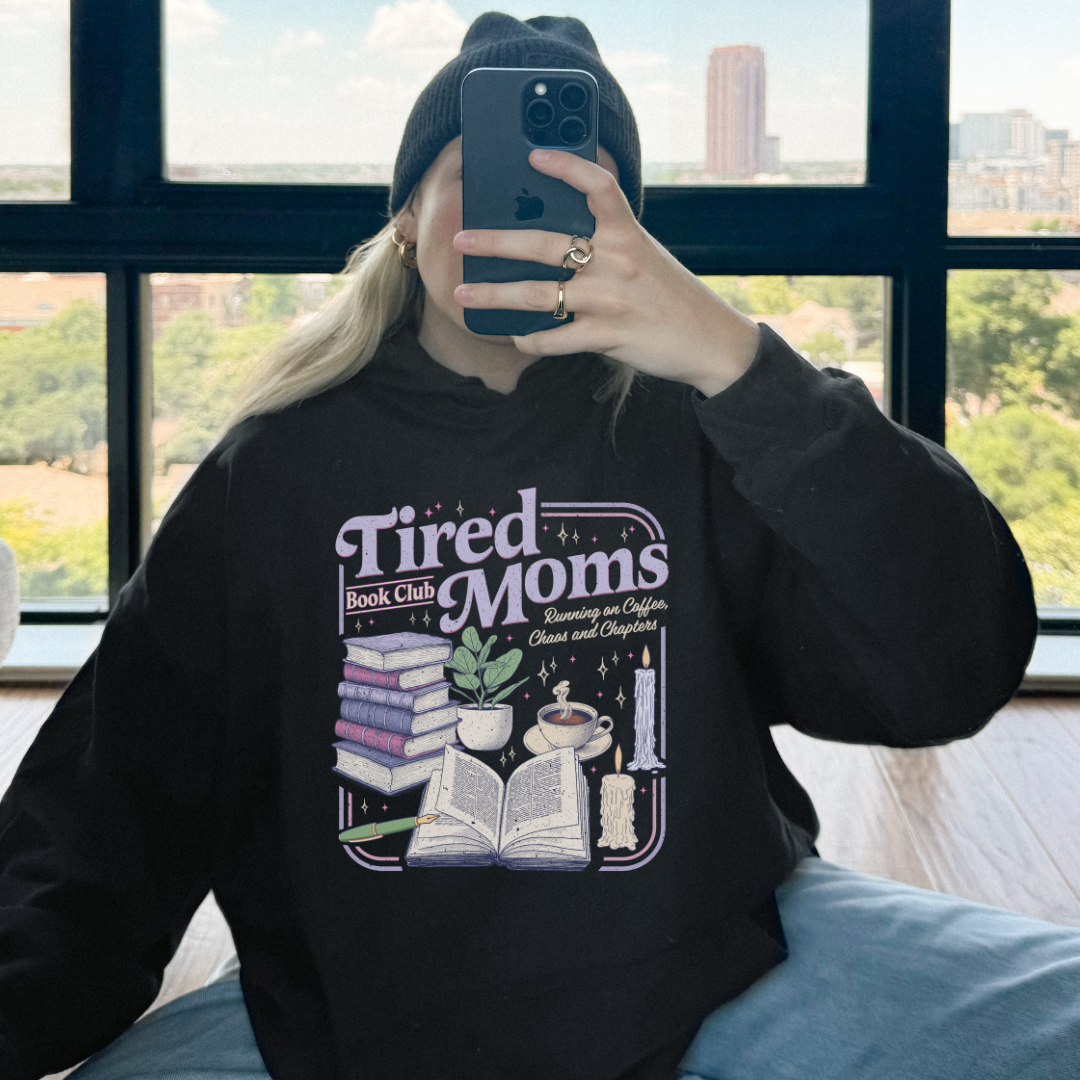 A person taking a mirror selfie wearing a black beanie and a black graphic hoodie with the text 'Tired Moms Book Club' and an illustration of books, a candle, and a plant. The background shows a large window with a city view.
