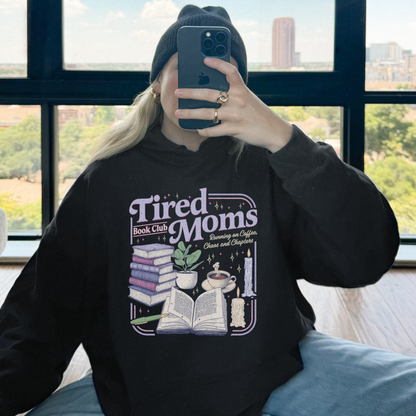A person taking a mirror selfie wearing a black beanie and a black graphic hoodie with the text 'Tired Moms Book Club' and an illustration of books, a candle, and a plant. The background shows a large window with a city view.