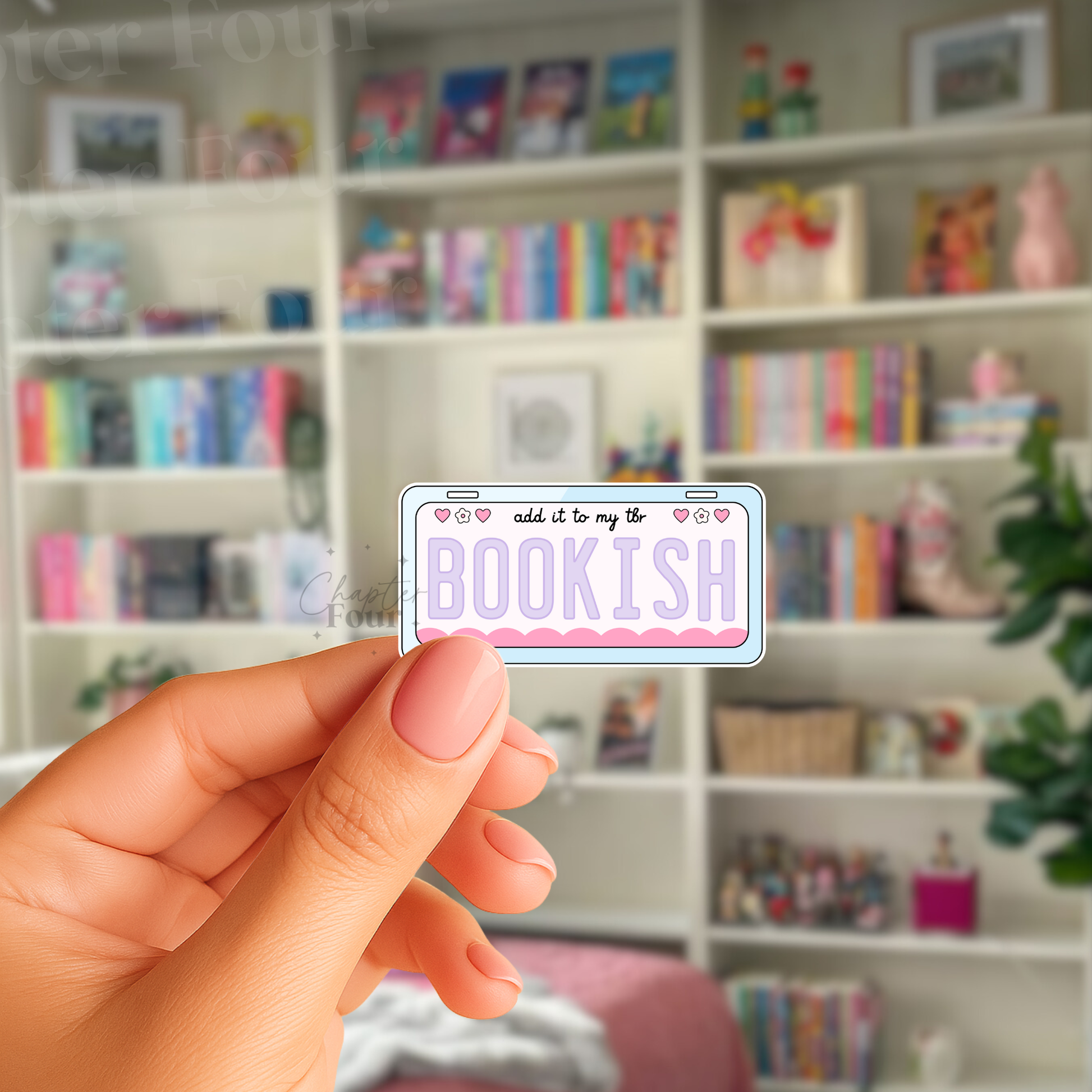 Hand holding a 'add it to my tbr BOOKISH'' license plate sticker in front of a bookshelf.
