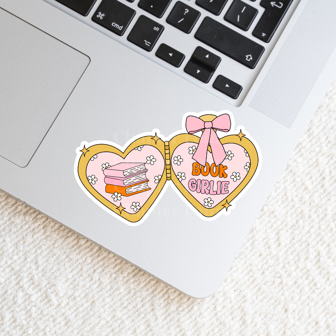 Heart-shaped locket sticker with 'Book Girlie' text on the right and 3 books stacked design on the left, laying on a laptop