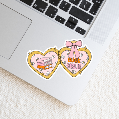Heart-shaped locket sticker with 'Book Girlie' text on the right and 3 books stacked design on the left, laying on a laptop