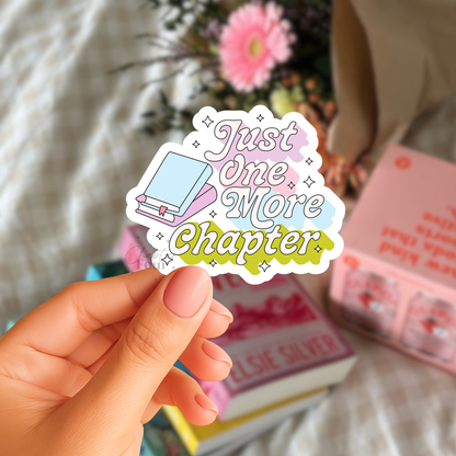Hand holding a 'Just One More Chapter' sticker with books and flowers in the background