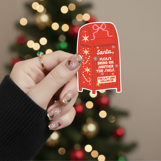 Hand holding a red Mail Drop box sticker with text that says Santa, Please Bring Me Another TBR Shelf The Last One Collapsed in front of a decorated tree.