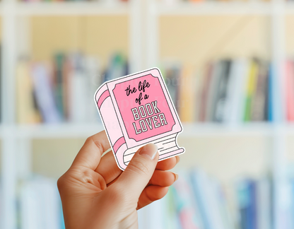 Hand holding a pink book-shaped sticker with 'the life of a book lover' text against a blurred bookshelf background.