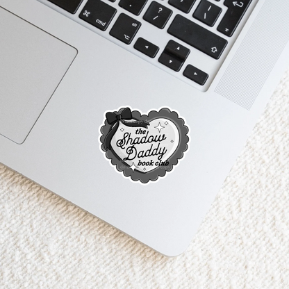 A gray and black heart sticker with the text 'The Shadow Daddy Book Club' placed on a laptop.