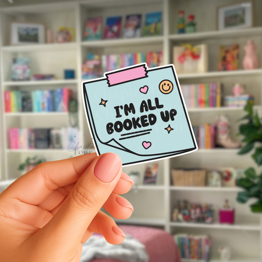 Hand holding a sticker with 'I'm all booked up' text in front of a bookshelf.