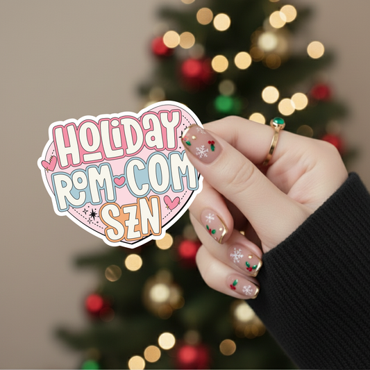 Hand holding a 'Holiday Rom-Com SZN' sticker with a Christmas tree in the background